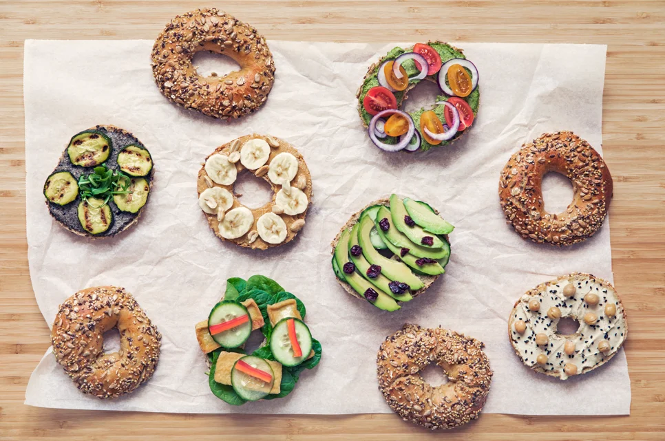 Assorted vegan bagels with plant-based toppings including avocado, tomato, and vegan cream cheese on a wooden board.