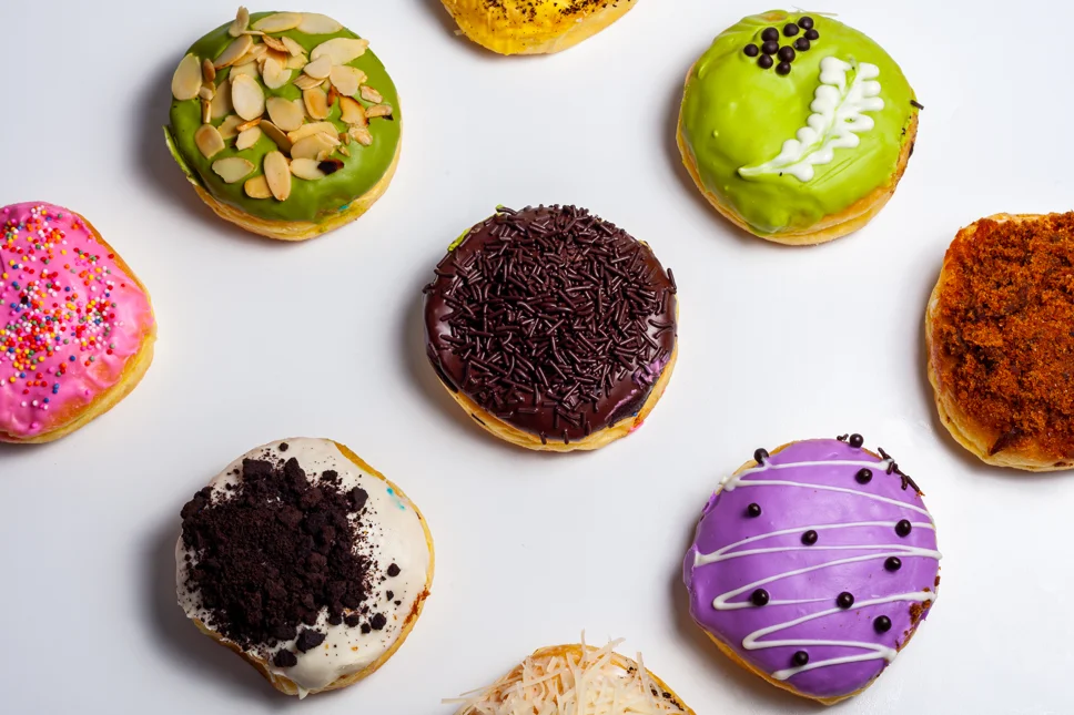 Assorted gourmet super donuts with vibrant toppings and unique flavors displayed on a wooden table, showcasing varieties from classic sugar-glazed to exotic matcha and bacon-infused creations.