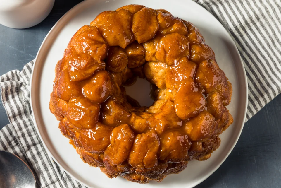 Freshly baked monkey bread on a serving plate, showing its golden caramelized crust and pull-apart texture, surrounded by cinnamon sticks and sugar for decoration.